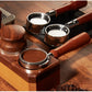 A wooden espresso preparation set with stainless steel portafilters, a tamper, and a press, arranged on a dark tray over a wooden surface.