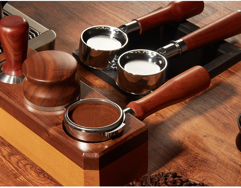A wooden espresso preparation set with stainless steel portafilters, a tamper, and a press, arranged on a dark tray over a wooden surface.