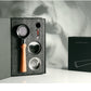 A coffee accessory set displayed in an open black presentation box, featuring a metal portafilter with a light wood handle, two metal filter baskets, and a small round tool. A closed black box with white line art stands beside it on a white surface against a gray background with soft shadow projections.