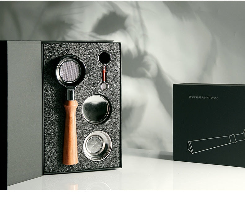 A coffee accessory set displayed in an open black presentation box, featuring a metal portafilter with a light wood handle, two metal filter baskets, and a small round tool. A closed black box with white line art stands beside it on a white surface against a gray background with soft shadow projections.