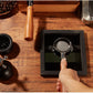 A hand holds a metal portafilter over a black square tray on a wooden tabletop, with coffee beans, a grinder, and other coffee-making tools arranged around it.