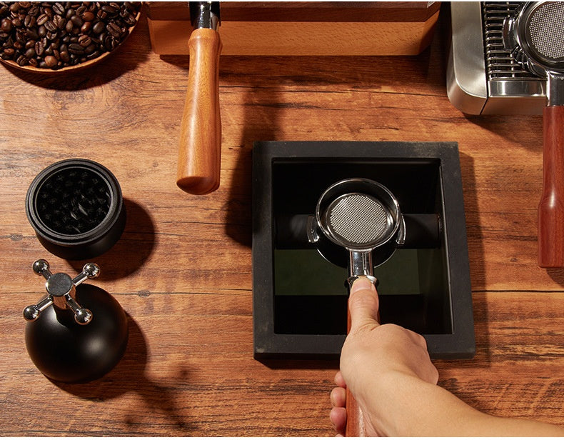 A hand holds a metal portafilter over a black square tray on a wooden tabletop, with coffee beans, a grinder, and other coffee-making tools arranged around it.